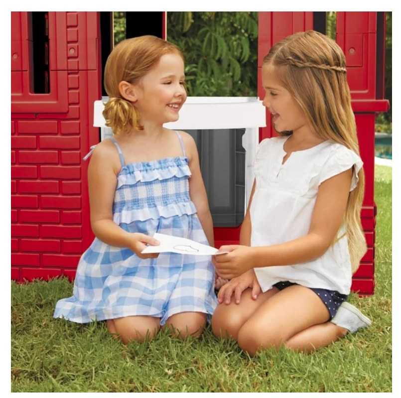 Two young girls sitting in front of a red playhouse, holding paper.