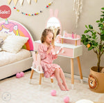 Young girl in a pink dress sitting on a chair in front of a small vanity table with a mirror, surrounded by toys and decor.